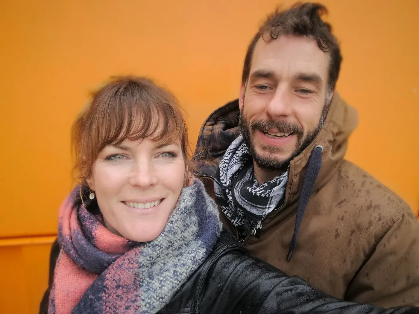 A smiling couple in scarves against an orange wall